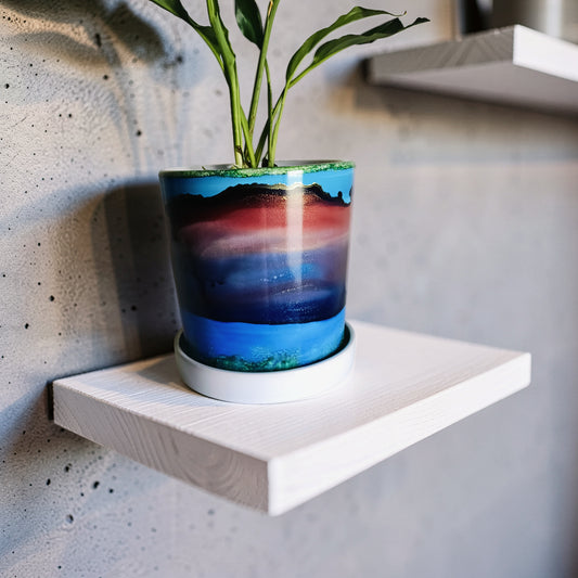 A cylindrical planter with a colorful landscape design resembling a sunset over water, sitting on a white wooden floating shelf against a textured gray concrete wall.