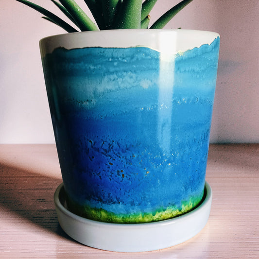 A cylindrical planter with a colorful landscape design resembling a sunset over water, sitting on a white wooden floating shelf against a textured gray concrete wall.