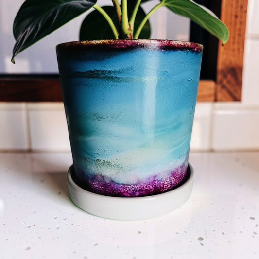 A glazed ceramic planter pot with a matching saucer, featuring a gradient design transitioning from teal blue to light blue and white, with a textured purple band at the bottom. A green plant is planted in the pot, set on a white tiled countertop.
