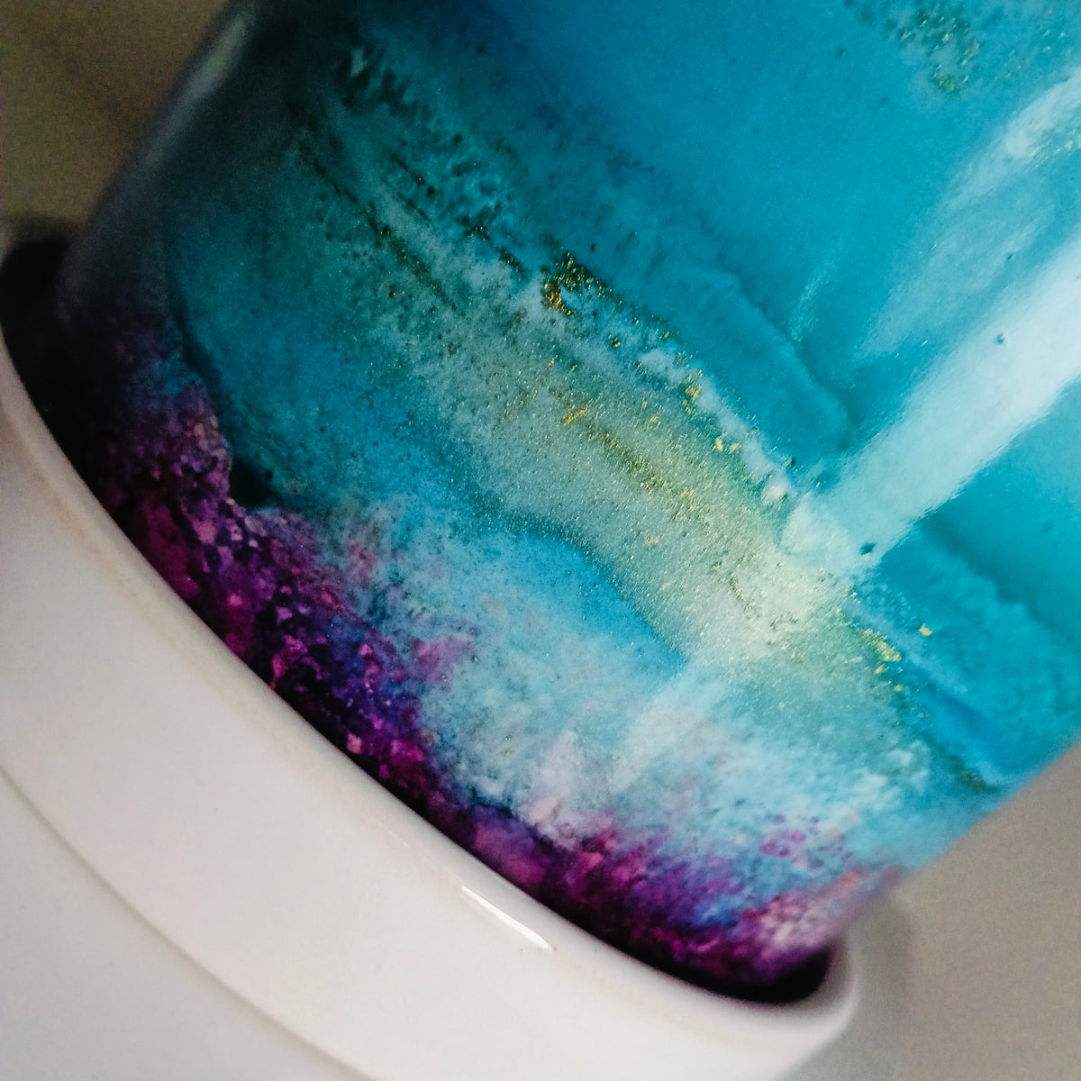 A close-up of a cylindrical product, likely a cup, tumbler, or container, featuring a marbled abstract design with vibrant turquoise blue, teal, purple, and white, accented with fine gold glitter. The product is shown resting on a white surface.