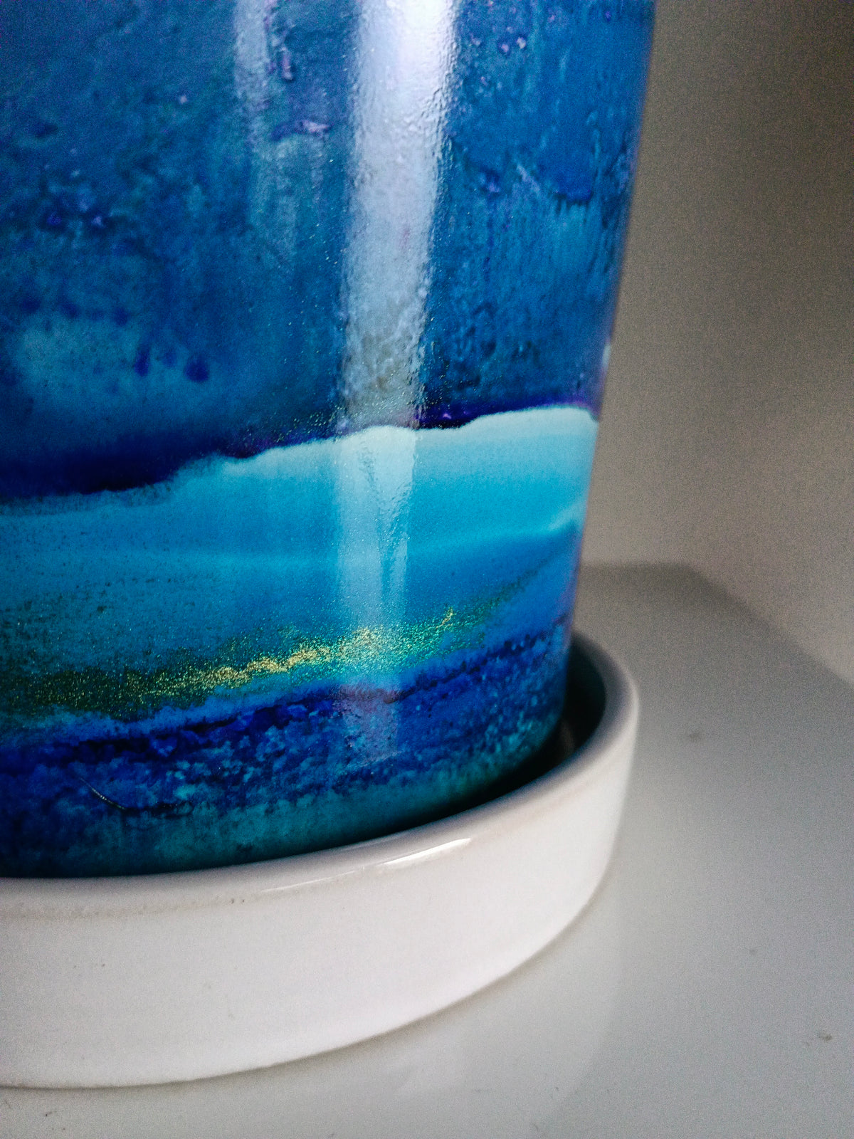 A close-up of a glossy ceramic cup or tumbler with a blue and turquoise landscape-style glaze, resting on a white saucer against a neutral light gray background.