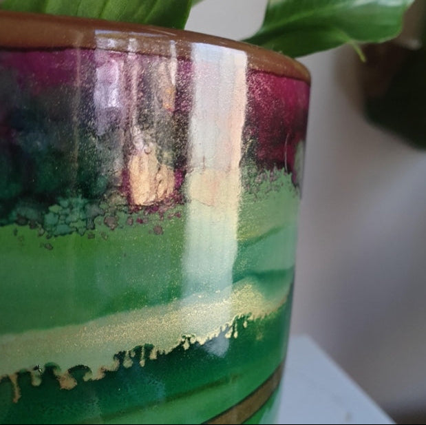 Close-up of a glossy ceramic planter with a marbled landscape design featuring green, purple, and gold glazes, with a plant leaf visible at the top.