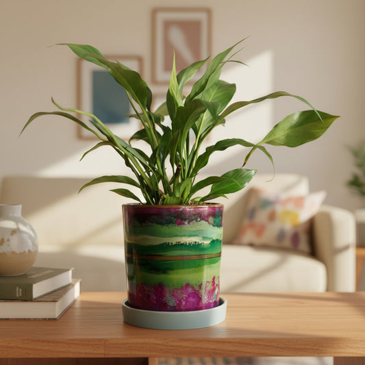 A potted peace lily plant with green leaves in a cylindrical ceramic pot featuring a colorful landscape design with green fields, purple mountains, and a gold band, sitting on a white saucer against a white background.
