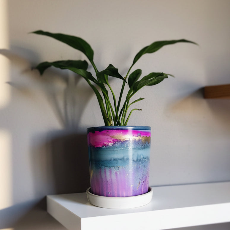A ceramic planter with a colorful marbled pattern in shades of purple, blue, and pink, holding a green plant, displayed on a white floating shelf against a gray wall.