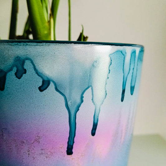 A close-up of a glazed ceramic planter with a blue, purple, and pink drip design, containing green plant stems against a light background.
