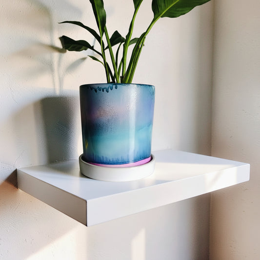 A glazed ceramic planter with a blue, purple, and teal gradient design, holding a green plant, displayed on a white floating shelf against a beige wall.