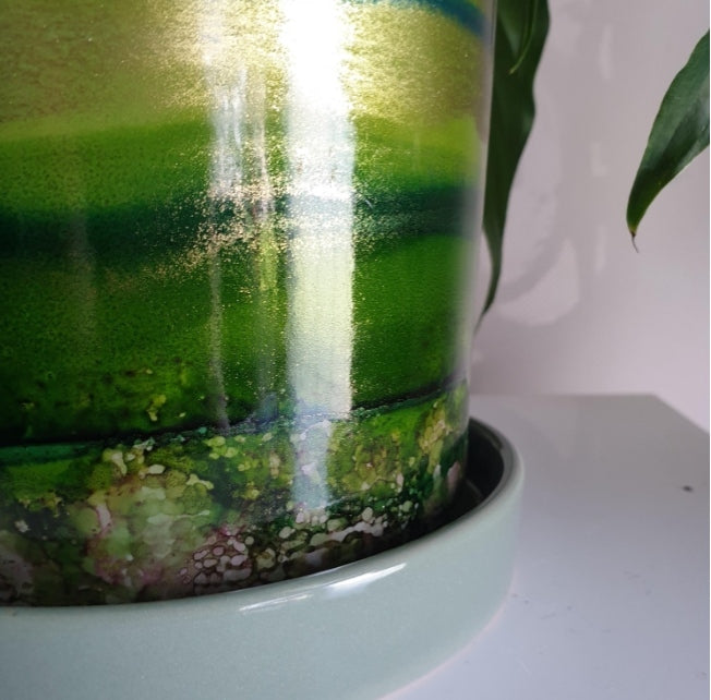 Close-up of a glossy ceramic planter with a green and gold marbled pattern, sitting on a white saucer with a plant leaf visible on the right.