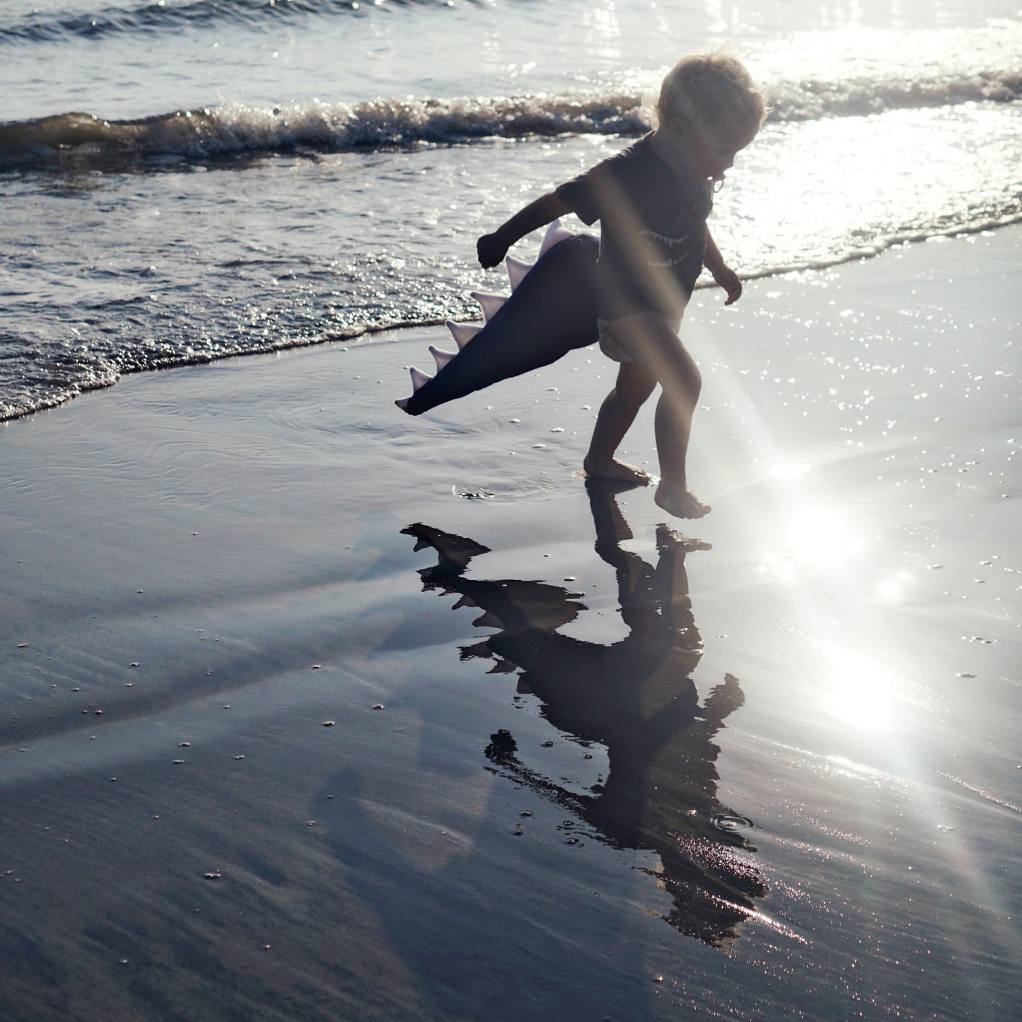 Child playing on a beach with waves in the background wearing a dino tail