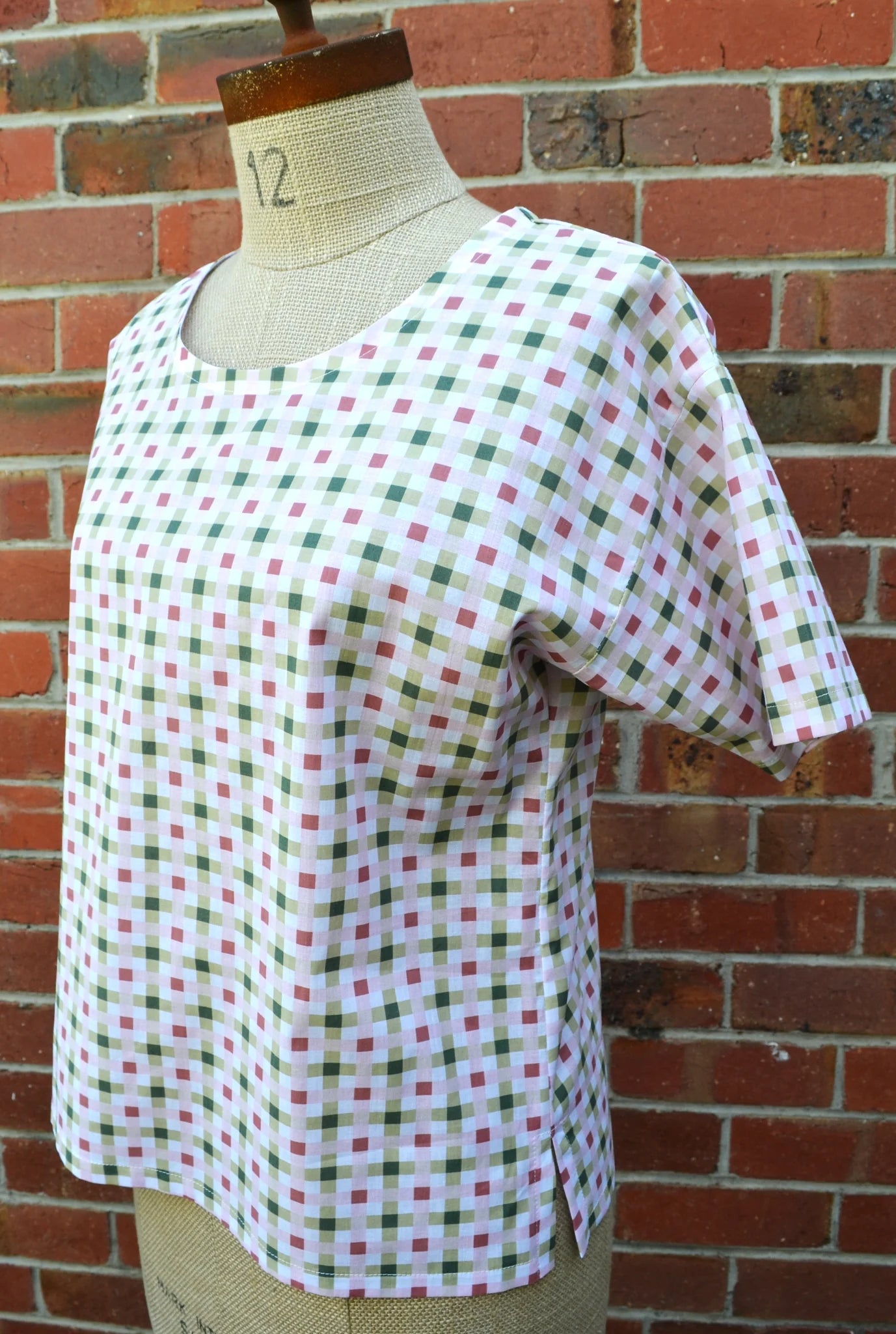 Mannequin wearing a pink and green checkered shirt against a brick wall.