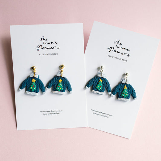 Pair of earrings resembling Christmas tree sweaters on branded cards against a pink background