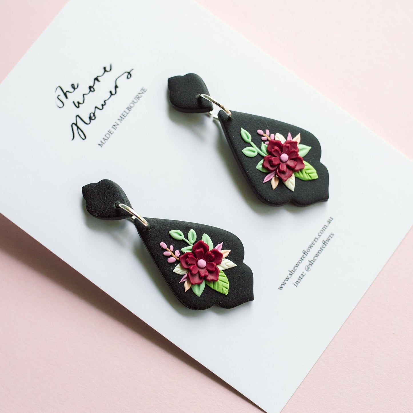 Black earrings with floral designs on a white card against a pink background