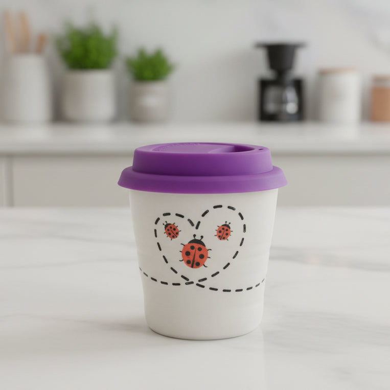A reusable white travel tumbler with a purple lid, decorated with a black dashed heart outline and three orange ladybugs, sitting on a wooden table in a shop.