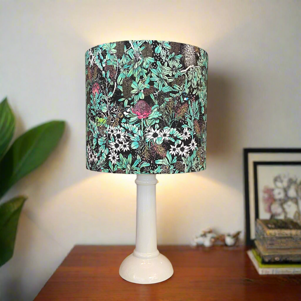 A White Mid Century Column Table lamp with a shade of native green flora and fauna sitting on a side table in a home