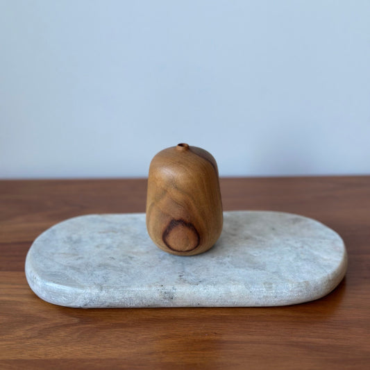 A small hand-carved wooden acorn-shaped object with a natural wood grain finish, displayed on a light gray stone tray over a wooden table against a light gray wall.