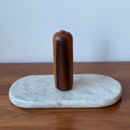 A dark brown wooden cylindrical object standing upright on a round light gray stone slab, set on a wooden tabletop against a pale gray wall.