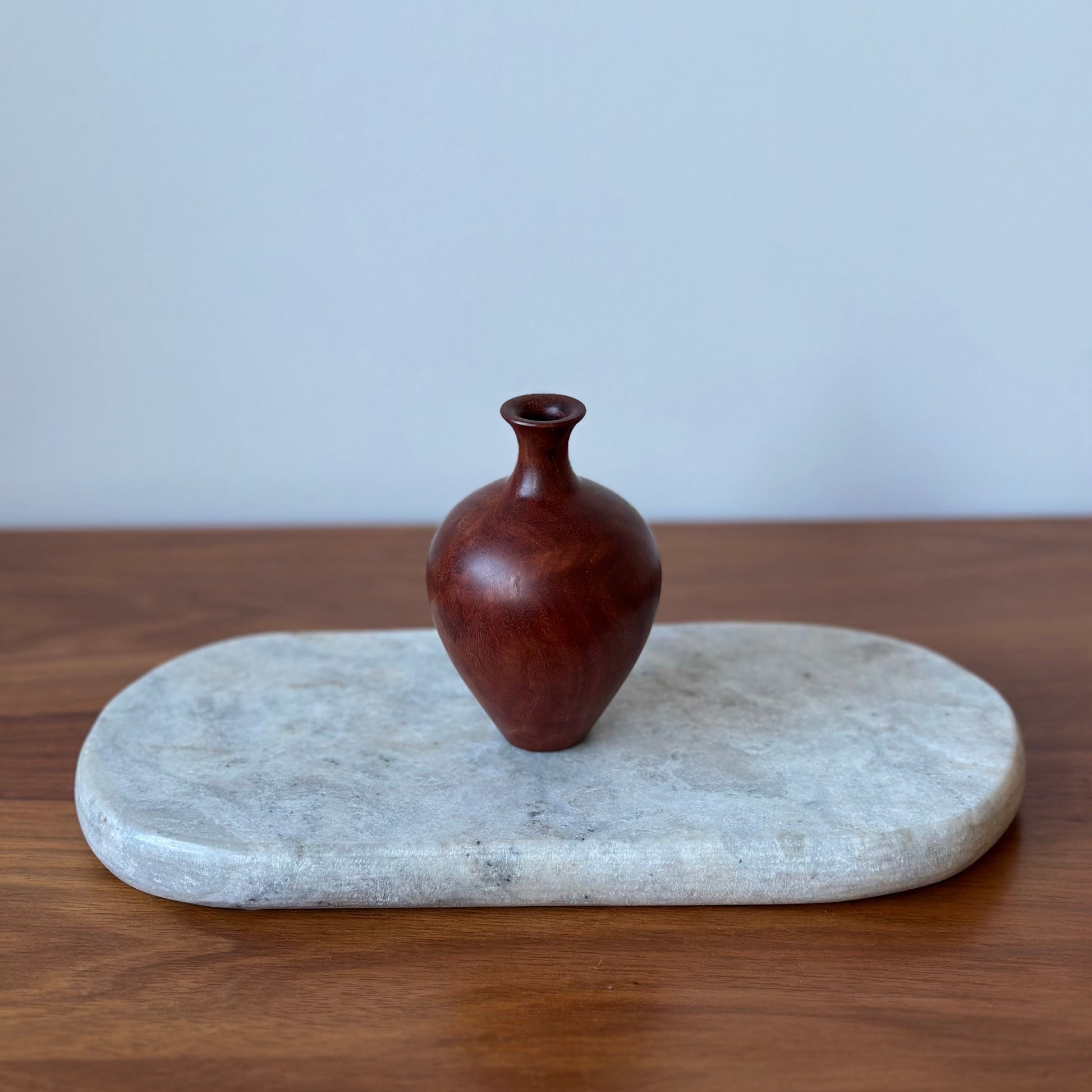 A small dark reddish-brown wooden vase with a bulbous body and narrow neck, displayed on a light gray stone tray over a wooden surface against a pale gray wall.