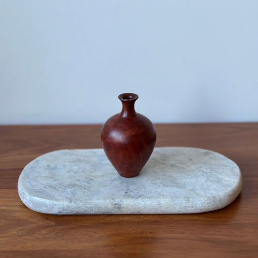 A small dark reddish-brown wooden vase with a bulbous body and narrow neck, displayed on a light gray stone tray over a wooden surface against a pale gray wall.