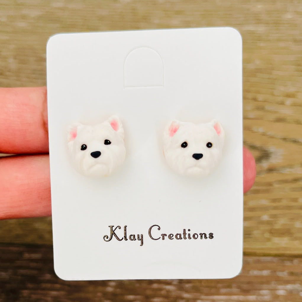 A pair of small stud earrings shaped like white dog faces with pink inner ears, displayed on a white backing card that reads 'Klay Creations' and held by a hand over a wooden surface.