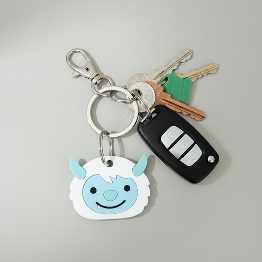 A handcrafted keyring featuring a smiling yeti character, made from laser cut acrylic and plywood, in blue and white colors, attached to stainless steel hardware.