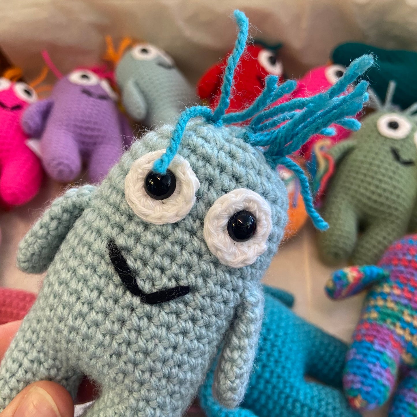 Close-up of a knitted toy with blue hair and black eyes, surrounded by other colorful knitted toys.