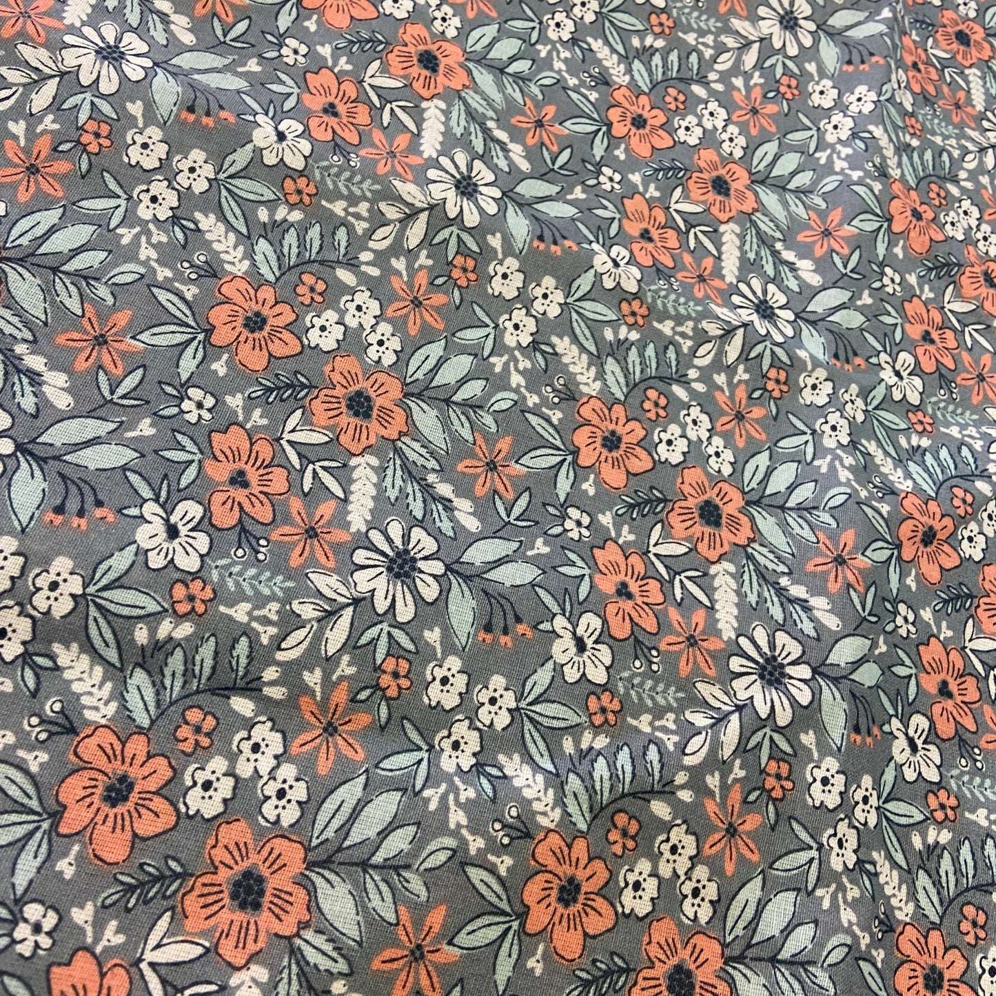 A close-up of a floral fabric pattern featuring orange, white, and light blue flowers with green leaves on a muted gray background.
