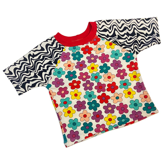 A short-sleeve raglan t-shirt laid flat on a light off-white background. The shirt features a cream-colored body covered in a retro-style multicolor flower pattern, a solid red crew neck, and black and white wavy patterned sleeves.