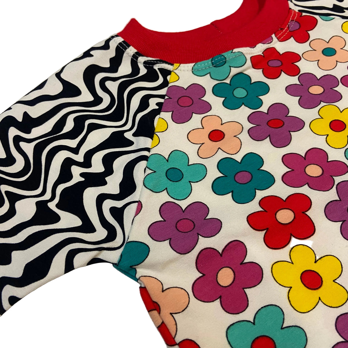 A close-up of a children's long-sleeve top with a split design: one sleeve and side panel have a black-and-white wavy zebra pattern, while the other side and front body feature a retro-style multicolor flower print on an off-white background with a red collar.