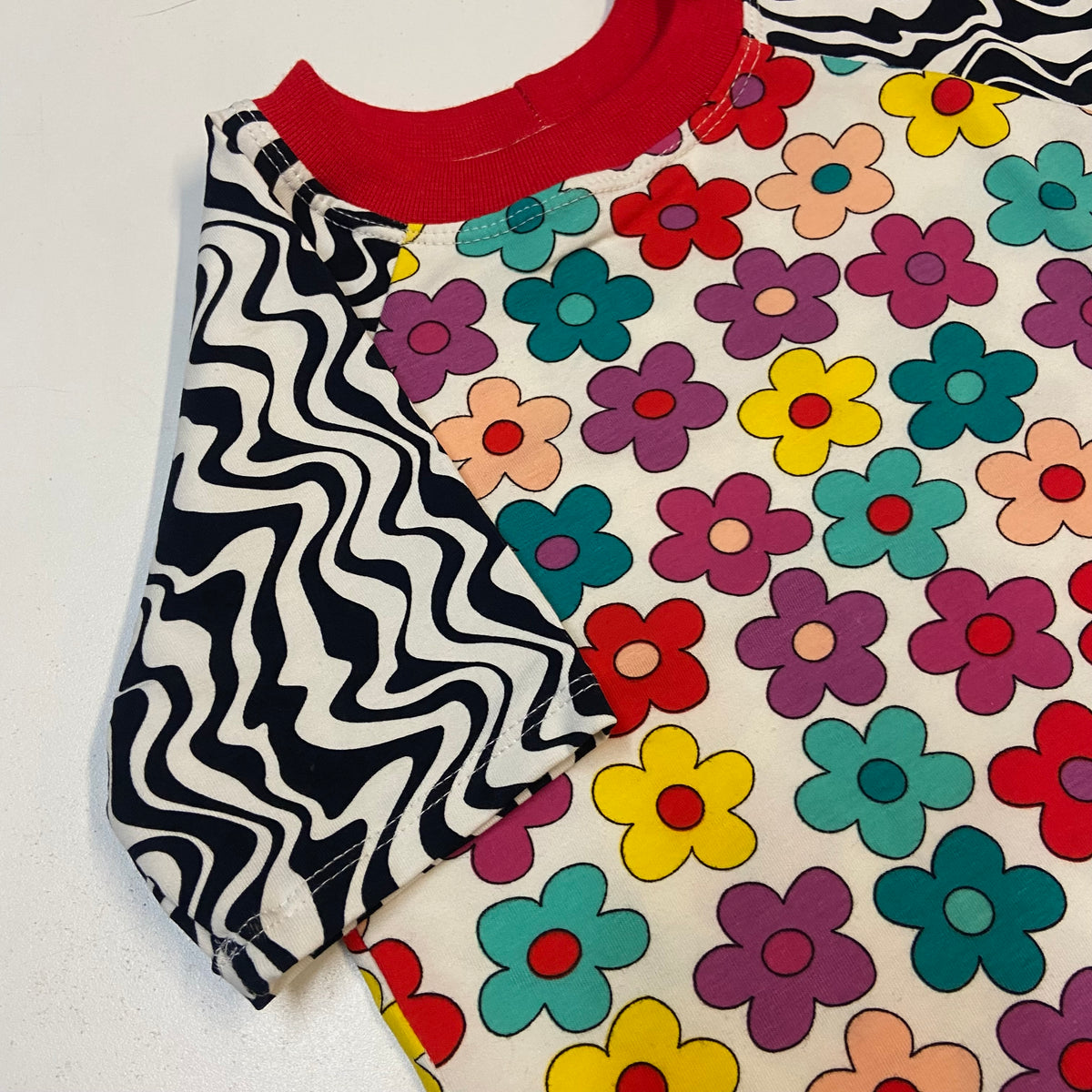 A close-up of a short-sleeve raglan shirt showing two contrasting fabric panels: one with a black and white wavy zebra-like pattern and one with a retro multi-colored flower print on a cream background, finished with a solid red ribbed crew neck.