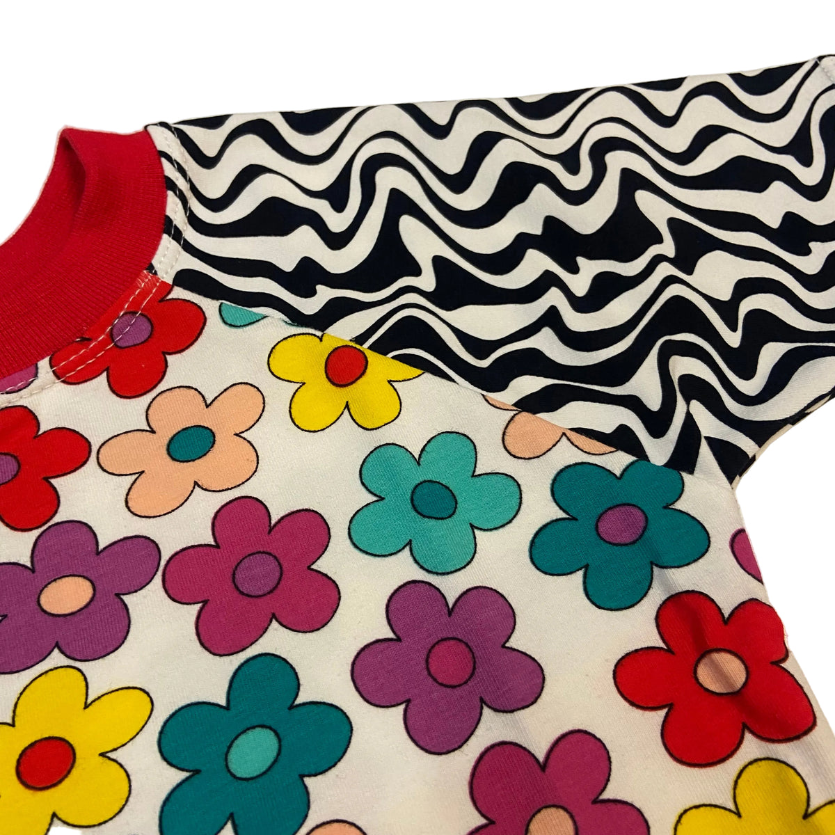 A close-up of a short-sleeve shirt laid flat on a light surface. The upper chest and right shoulder area show a black-and-white wavy stripe pattern, while the lower body is white with large colorful flowers in red, yellow, teal, purple, and peach.