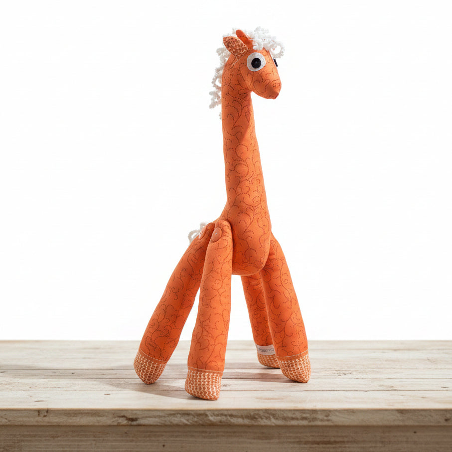 An orange plush giraffe toy with a patterned body, white textured mane and tail, and large black-and-white eyes, standing on a gray floor against a gray tiled wall.