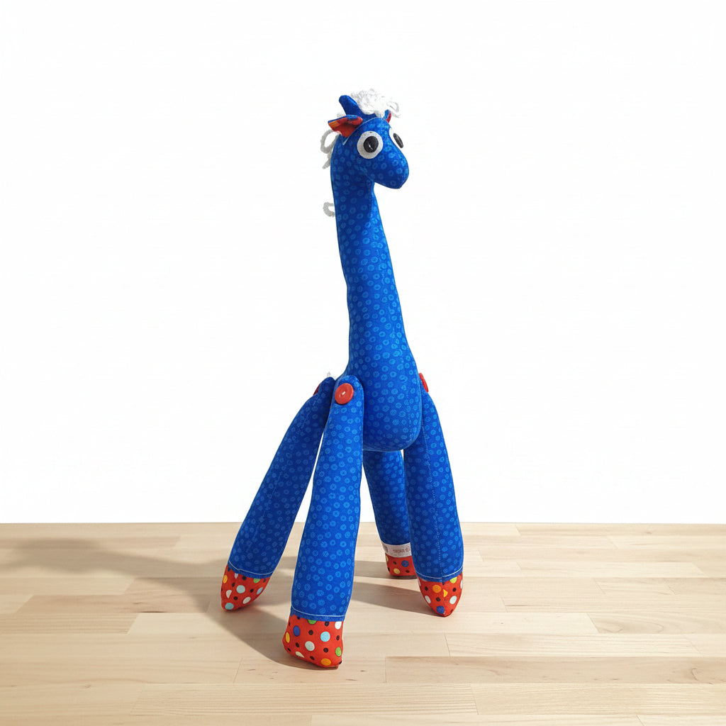 A handmade blue giraffe plush toy with white polka dots, red and white polka-dotted hooves, and a white mane, standing on a gray tiled floor against a gray tiled wall.