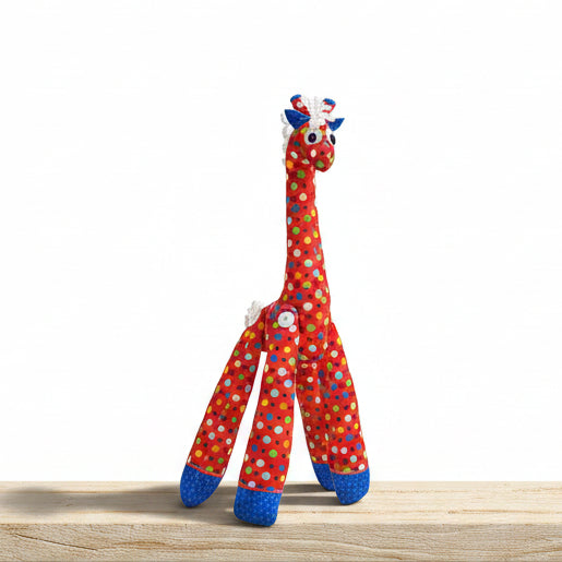 A handmade fabric giraffe toy standing on a gray tiled floor against a gray tiled wall. The giraffe is primarily red with multicolored polka dots (blue, green, yellow, white) and has blue hooves and a white mane.