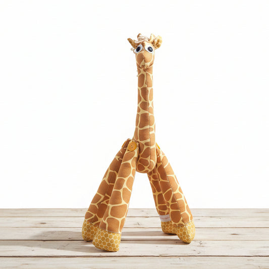 A plush giraffe toy standing on a gray floor against a dark gray tiled wall. The giraffe is tan and brown with white spots, large cartoon eyes, and a small yellow button on its neck.