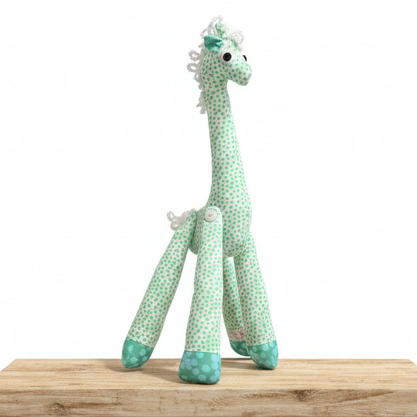 A handmade plush giraffe toy with a mint green and white spotted pattern, featuring a white lace mane and tail, standing on a gray textured floor against a dark gray tiled wall.
