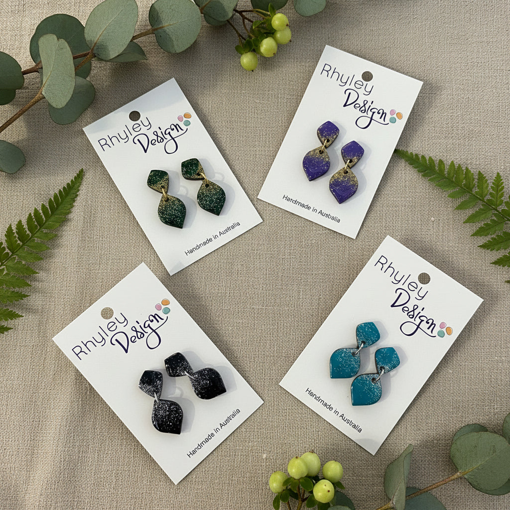 Four pairs of Rhyley Design statement earrings displayed on white backing cards on a wooden surface. The earrings feature organic, wavy shapes with a glittery finish in green, purple, black, and teal blue.
