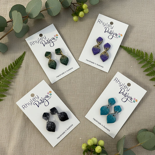 Four pairs of Rhyley Design statement earrings displayed on white backing cards on a wooden surface. The earrings feature organic, wavy shapes with a glittery finish in green, purple, black, and teal blue.