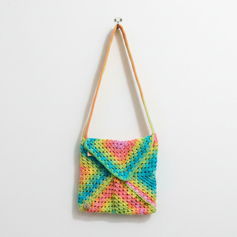 A crocheted shoulder bag with a vibrant multicolor neon pattern of pink, yellow, green, and blue, resting on a textured concrete floor with a person's red sneakers visible at the bottom edge.