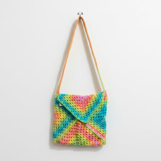A crocheted shoulder bag with a vibrant multicolor neon pattern of pink, yellow, green, and blue, resting on a textured concrete floor with a person's red sneakers visible at the bottom edge.