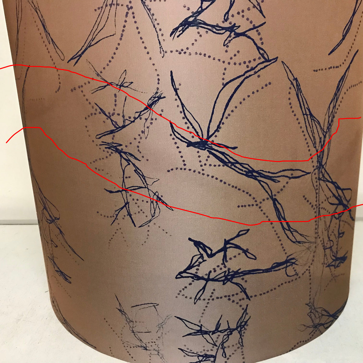 A close-up of a cylindrical lampshade or drum shade in warm taupe-beige with a dark navy abstract line and dotted pattern across the surface.