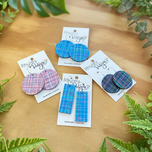 Four pairs of handmade plaid earrings on white display cards, arranged on a light wood surface. The earrings feature blue, pink, and green tartan patterns in various shapes including circles, rectangles, and a mix of both.