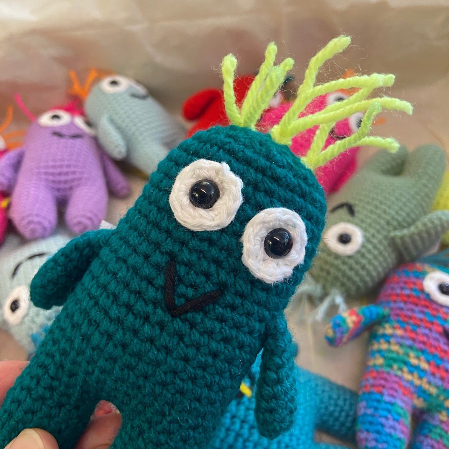 Handmade jade crochet creature with large eyes and a small mouth, surrounded by other colorful crochet toys.