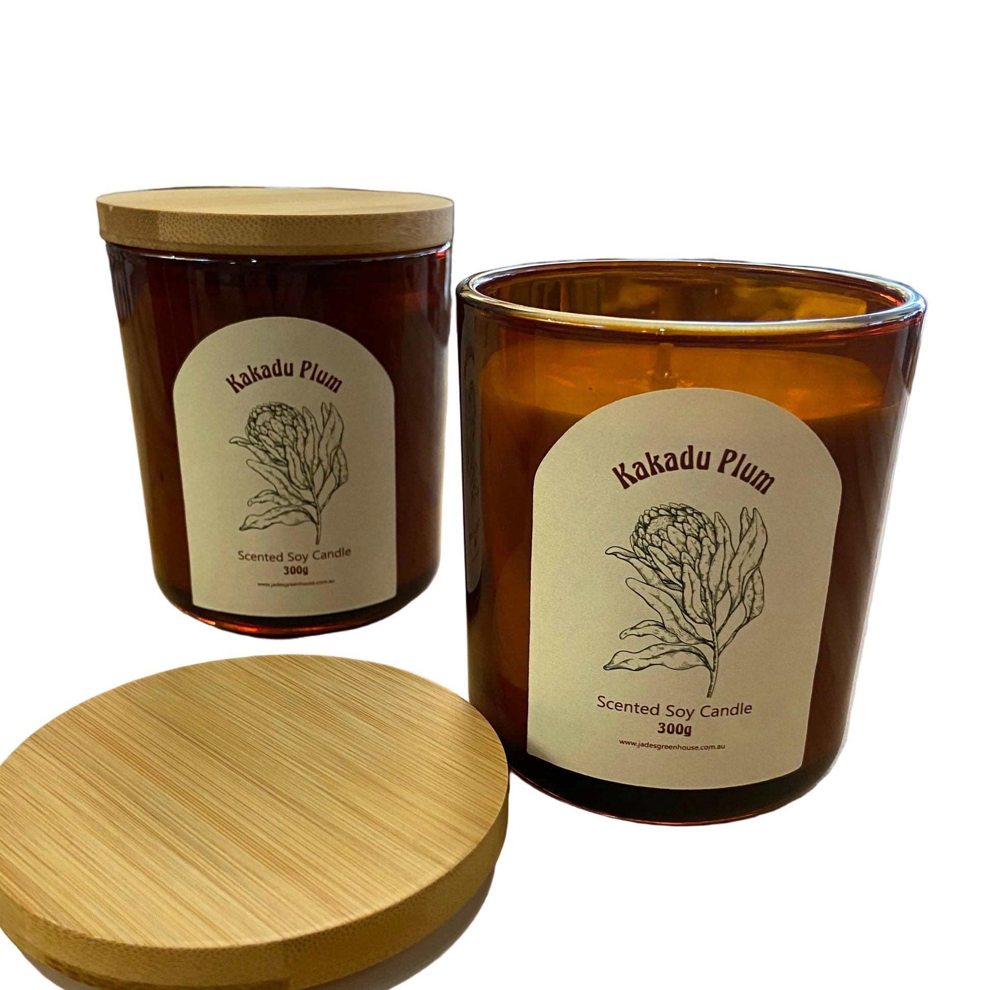 Two amber glass scented soy candles with bamboo lids on a wooden table inside a shop. The front candle is open, showing the wax, and the label reads 'Akadu Plum'.