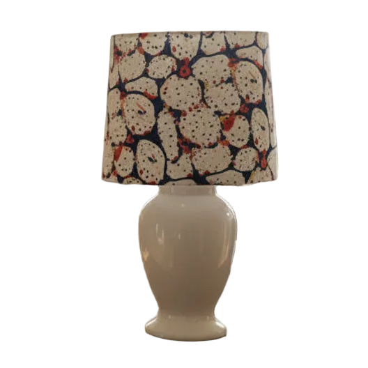 A table lamp with a glossy white ceramic base and a patterned drum shade featuring a cream background with dark blue and red abstract spots, styled on a white background
