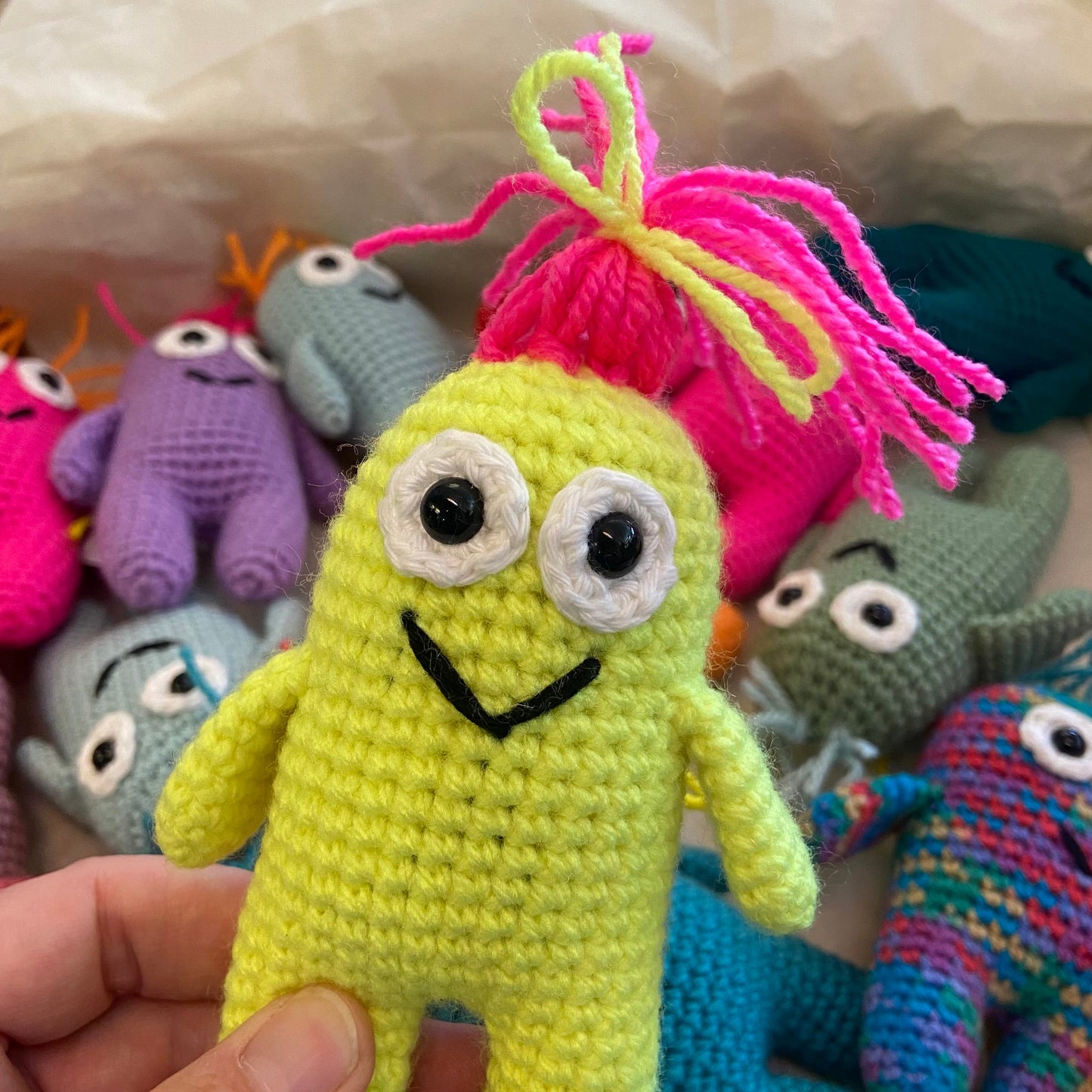 Hand holding a neon green crochet toy with googly eyes and pink hair, surrounded by other colorful crochet toys.