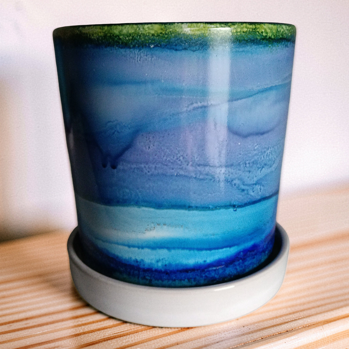 A glazed ceramic cup with a blue and purple marbled pattern, featuring a green rim and resting on a white saucer, set on a wooden surface against a neutral background.