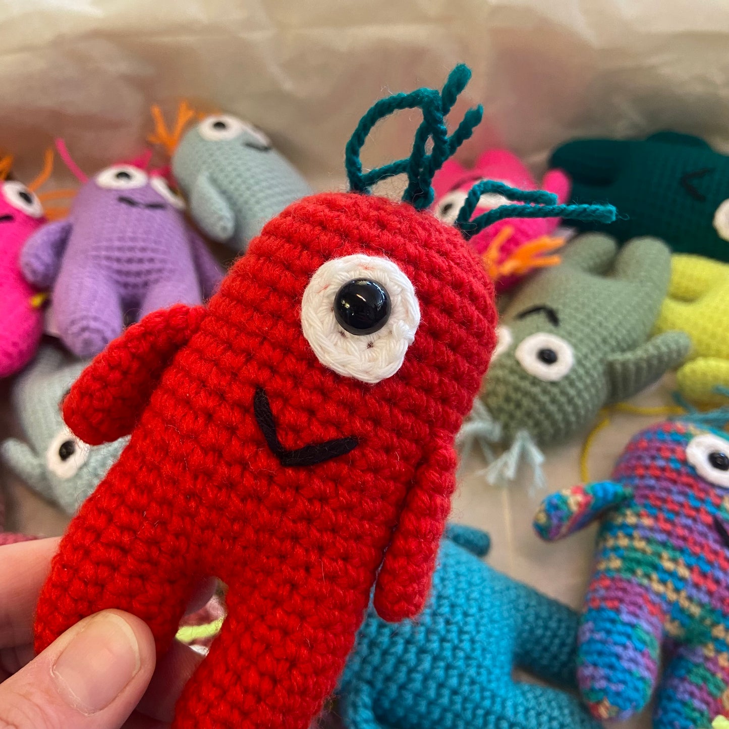 Hand holding a red crochet toy with multiple colorful toys in the background.