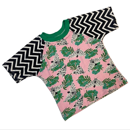 A short-sleeve raglan t-shirt laid flat on a light beige background. The shirt features a pink body with a green and black leopard print, black and white chevron-patterned sleeves, and a solid green collar.
